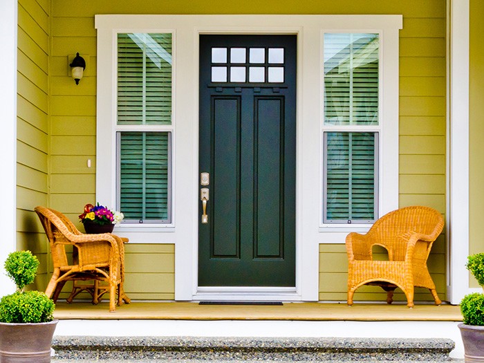 Yellow wooden home with gorgeous curb appeal and two woven chairs and a black door.